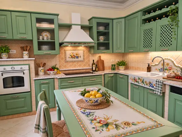Cucina angolare Siracusa con ante verde salvia, forno in colonna e tavolo con decori di frutta e fiori