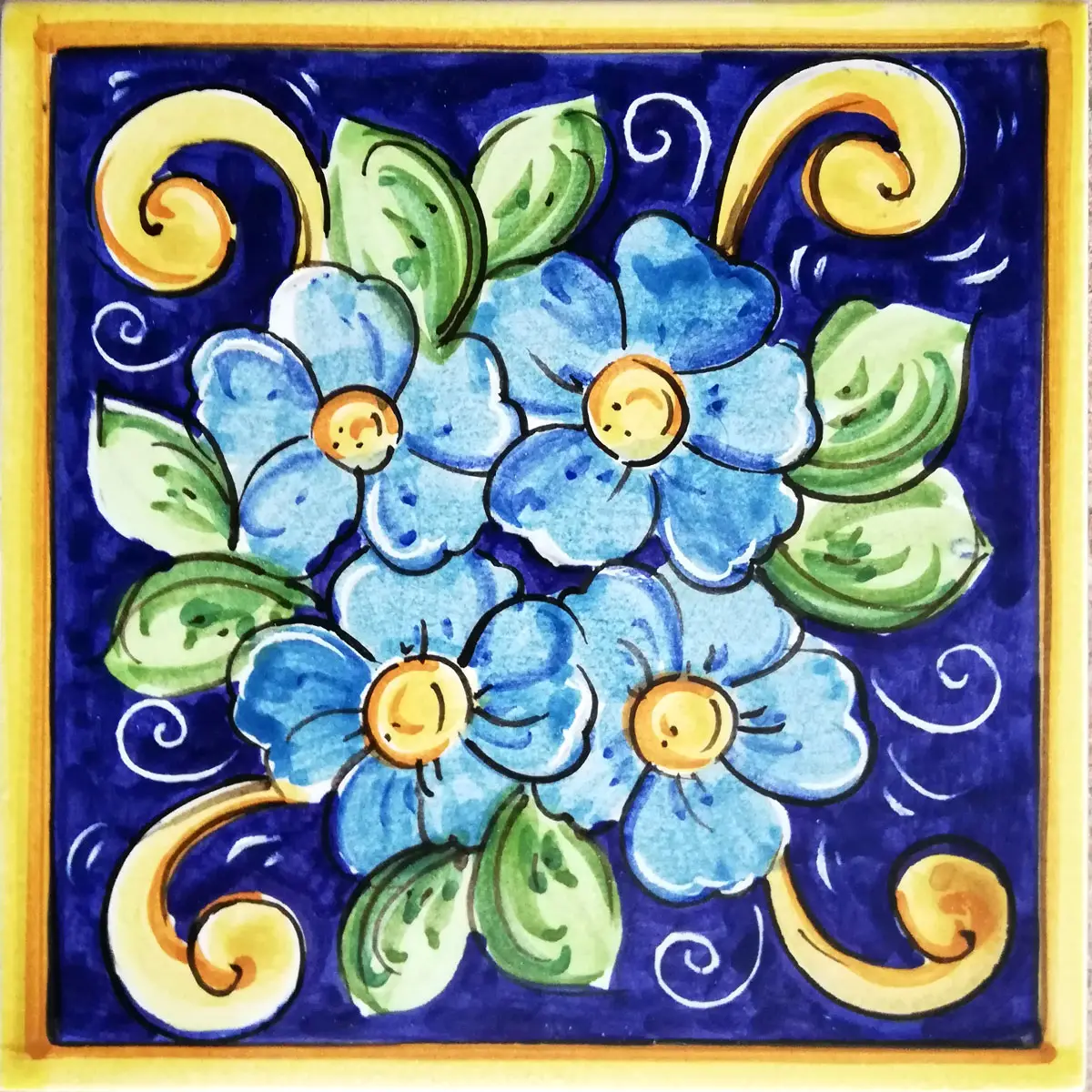 Mattonella decorata con fiori azzurri, foglie verdi e volute gialle su fondo blu con bordo giallo